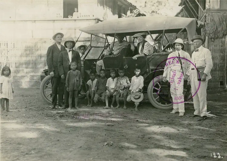 Hapag (HAPAG) tourists posing in and around a car with a local driver and children in front of traditional houses.